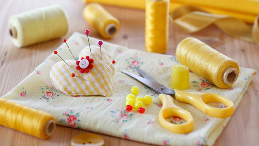 Sewing supplies for beginners, including yellow fabric and thread, scissors, and pins stuck in a heart-shaped cushion.