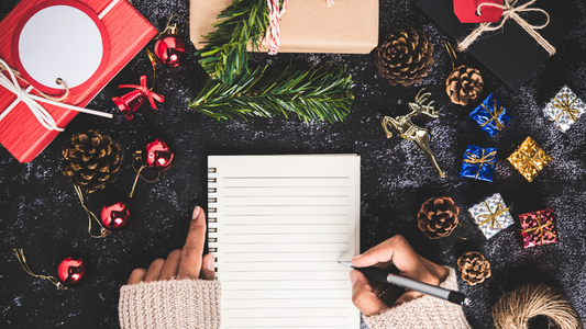 A tabletop covered with presents and ornaments features an empty notebook page and the hands of a person who is about to start planning their homemade holiday gifts.