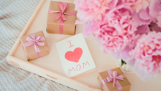 Pink flowers with three wrapped simple Mother’s Day gifts and a card that says “I (heart) mom.”